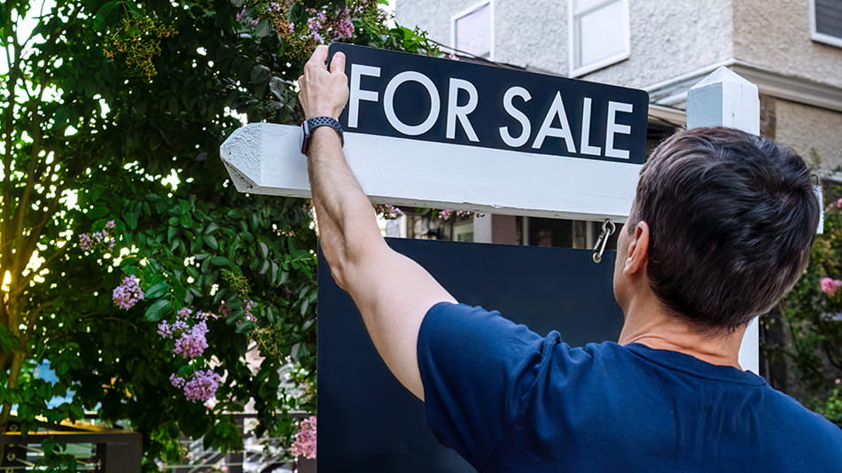 Persona colocando un cartel de venta frente a su casa porque los precios de las viviendas y las tasas de interés podrían no bajar en el corto plazo.